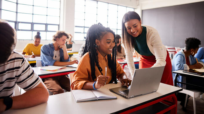 Smiling young female teacher helping a student working on a laptop during a lesson in a school classroom
