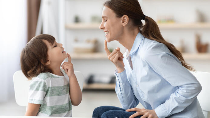 Speech training for kids. Professional woman specialist training with little boy at cabinet, teaching him right articulation exercises, side view