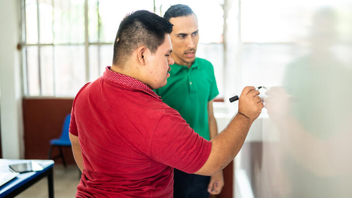Young man with Down Syndrome is solving a math problem while his male teacher, who has a cognitive disability, looks on.