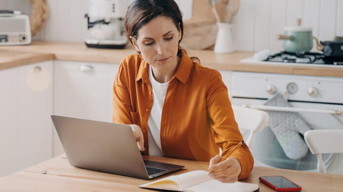 woman in orange shirt on her laptop taking notes in her kitchen