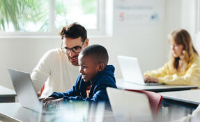Male teacher helps a young boy with computer-based learning in a classroom setting. Child tutor providing a lesson in an elementary school, with a focus on coding and basic digital literacy.