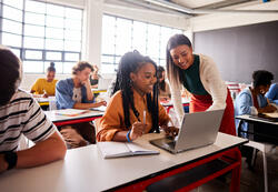 Smiling young female teacher helping a student working on a laptop during a lesson in a school classroom