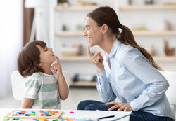Speech training for kids. Professional woman specialist training with little boy at cabinet, teaching him right articulation exercises, side view