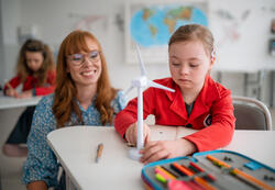 A down syndrome schoolgirl with model of wind turbine with help of teacher learning about eco-friendly renewable sources of energy in class at school, integration concept.
