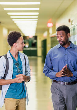 An African-American high school student walking with his teacher or school principal, in the hallway by a row of lockers. They are talking. The teacher has a serious expression on his face, a mature man in his 50s. The male student is an 18 year old teenage boy carrying a backpack.